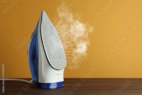 Modern iron with steam on wooden table against orange background, space for text