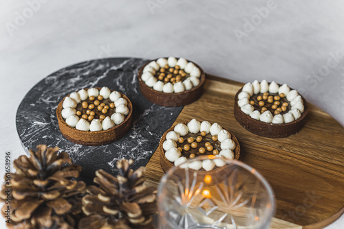 Close-up shot of four mini chocolate dessert tarts with golden sprinkles on wood and marble tray with lit candle and decorative pinecones. Festive baking. Horizontal shot. High quality photo