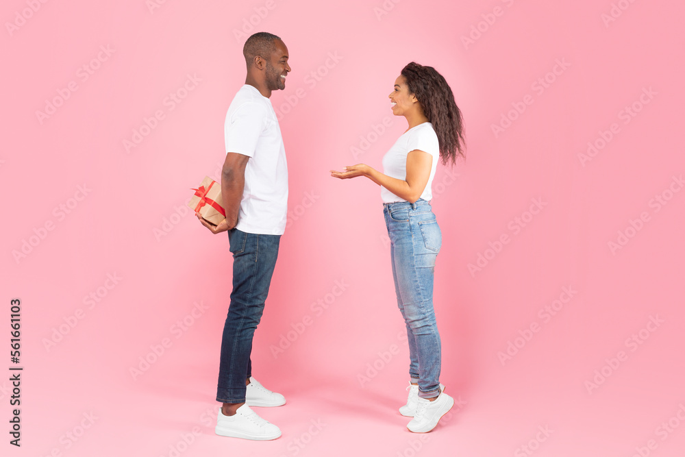 African american middle aged man hiding gift box behind back and looking at happy interested woman, pink background African american middle aged man hiding gift box behind back and looking at happy interested woman, pink background