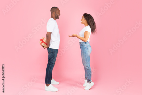 African american middle aged man hiding gift box behind back and looking at happy interested woman, pink background