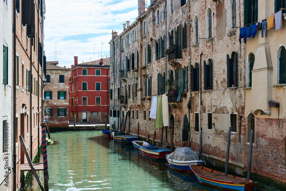 Picturesque facades of old buildings with hanging clothes and boats parked on canal in Venice, Italy, under blue sky with clouds on spring day. Beautiful view in famous Italian landmark.