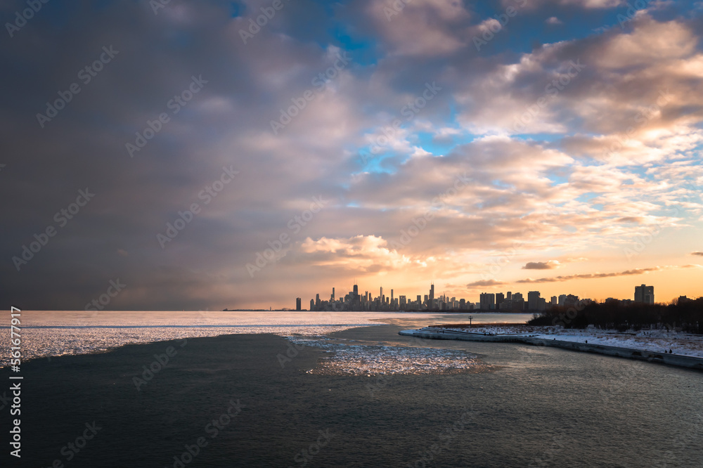 Aerial view of downtown Chicago Skyline from above the icy cold water of Lake Michigan in winter as the sunset colors the blue sky and clouds with orange and yellow hues.
