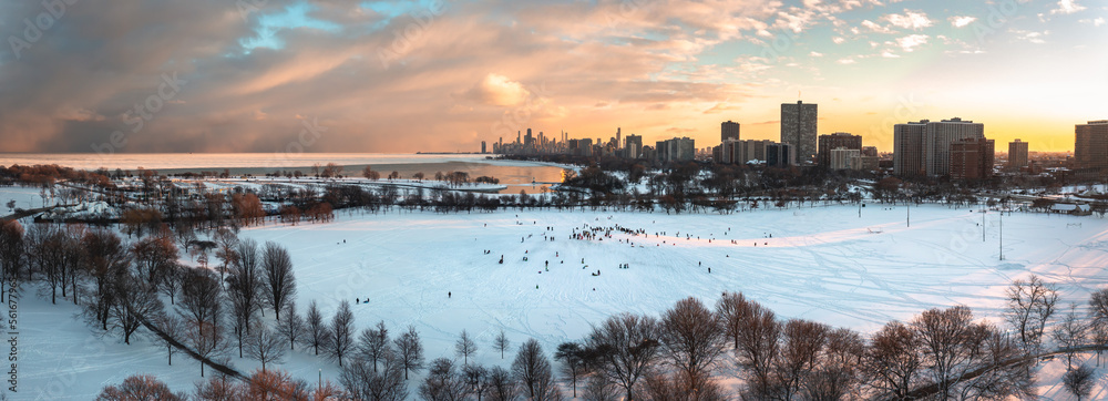 Wide angle aerial panorama of people sledding down Cricket Hill at sunset with bare trees and snow below and a bright yellow and pink sunset over the Chicago skyline beyond.