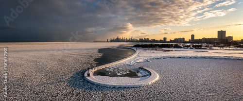 Beautiful Chicago skyline aerial drone view from above the frozen ice and snow covering Lake Michigan with a curved hooked pier near Montrose beach with colorful orange and pink sunset clouds above.