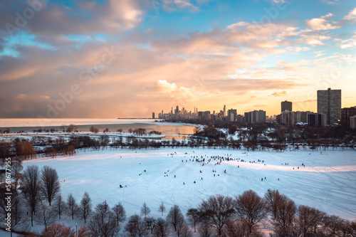 Crowds of people and families gather at Cricket hill to go sledding on a winter evening as the sunset begins to light up the clouds and sky in hues of yellow and pink.