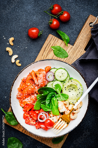 Ketogenic diet salad bowl with salmon, shrimp, avocado, spinach, cucumber, tomato, cashew nuts, sesame. Low-carbohydrate breakfast rich in healthy fats. Black table background, top view