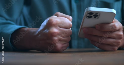 Close-up stressed man uses smartphone and loses his temper reading bad news on the internet, hits his fist on the table. Stressed male violently slams his fist on the table, showing anger.