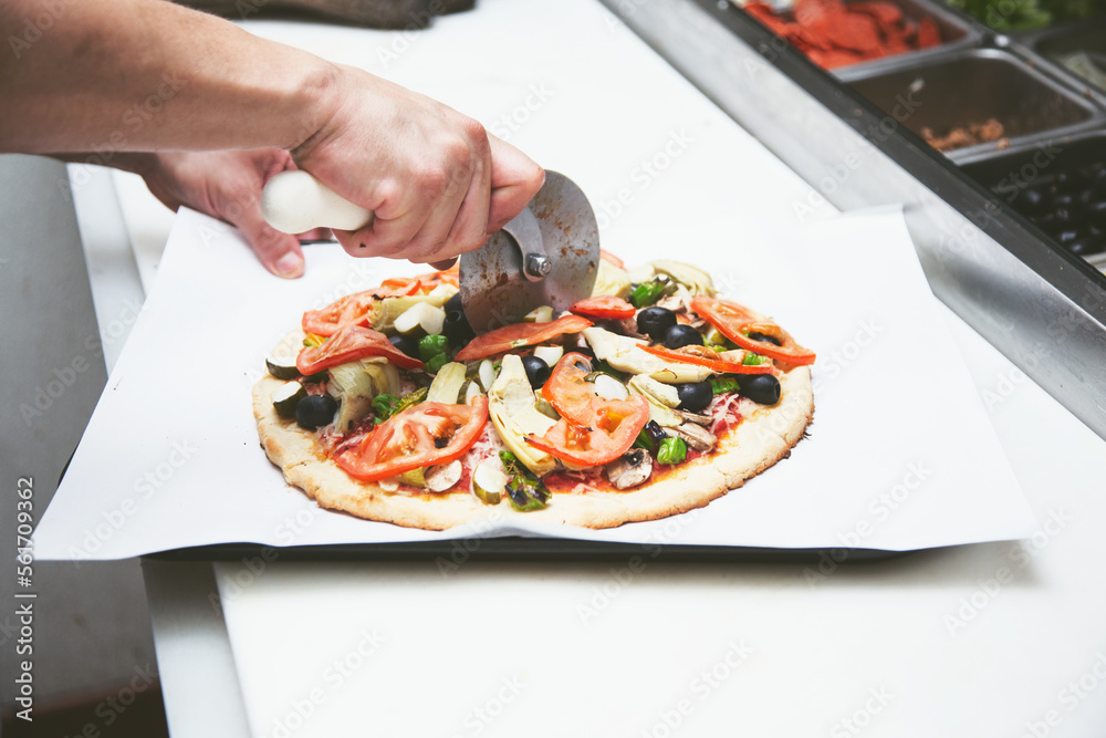A view of a vegan pizza at an ingredient station. The cook uses a pizza slicer to produce slices.
