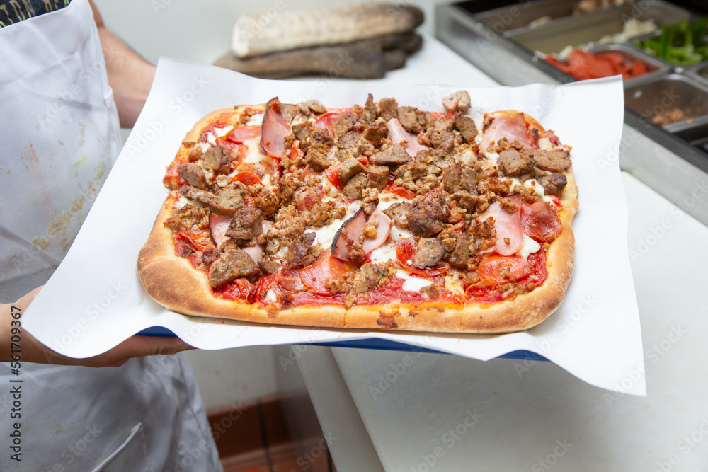 A view of a cook holding up a meat lovers pizza, seen at a local pizzeria.