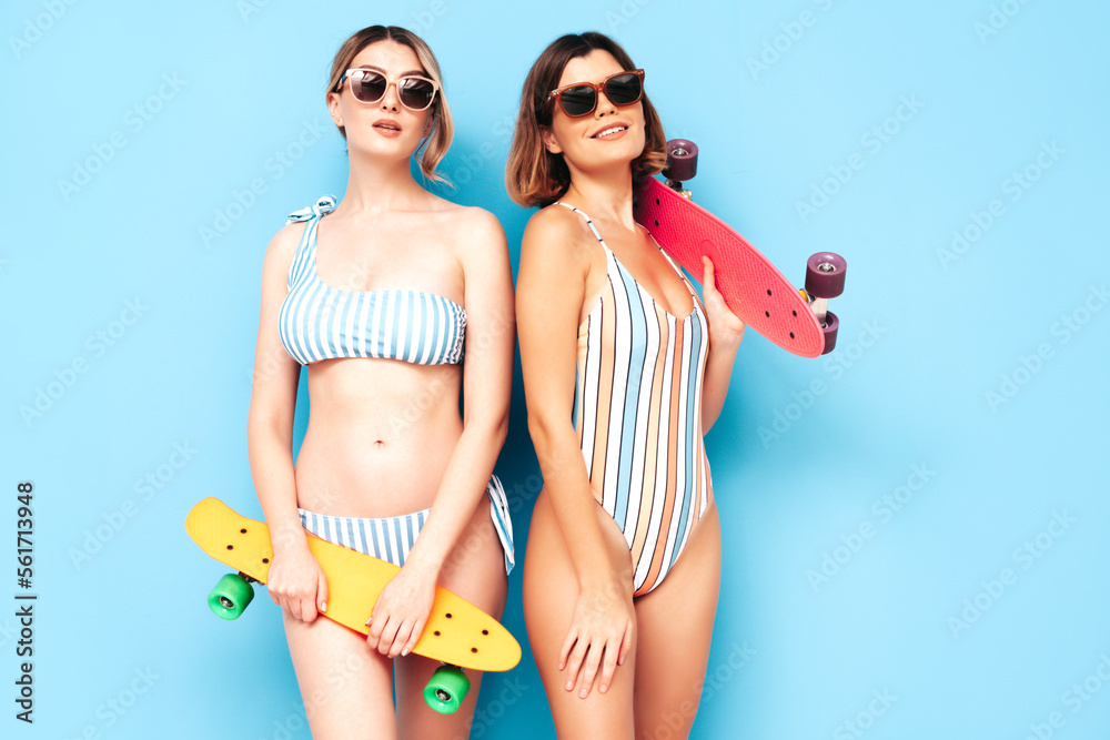 Two beautiful sexy smiling hipster women in summer swimwear bathing suits. Trendy models having fun in studio. Hot female isolated on blue. Cheerful and happy. In sunglasses. Holding penny skateboard Two beautiful sexy smiling hipster women in summer swimwear bathing suits. Trendy models having fun in studio. Hot female isolated on blue. Cheerful and happy. In sunglasses. Holding penny skateboard