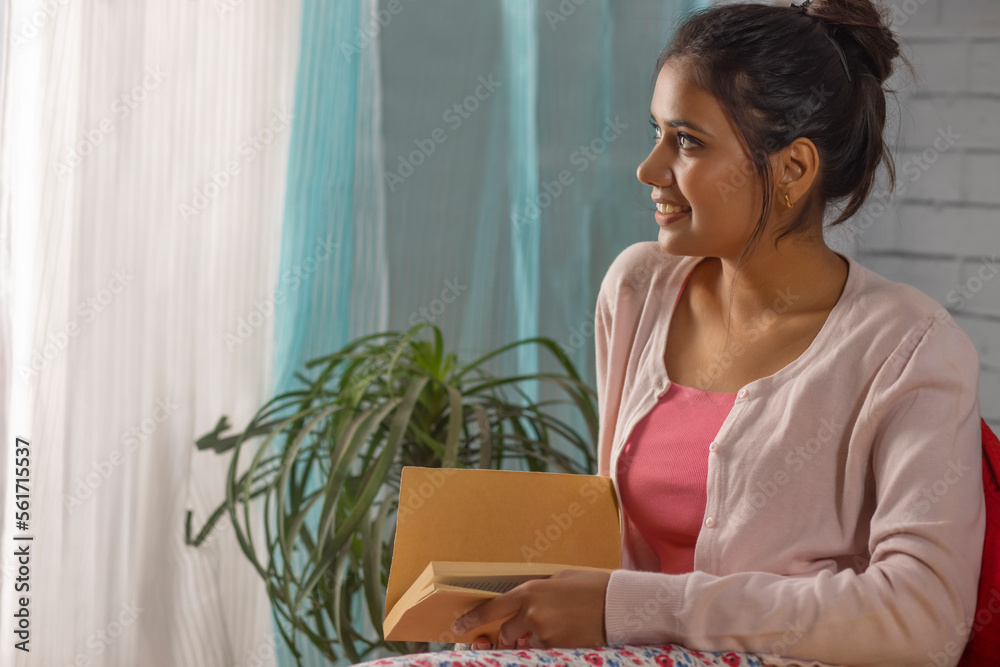 Young woman looking at window while reading book in living room Young woman looking at window while reading book in living room