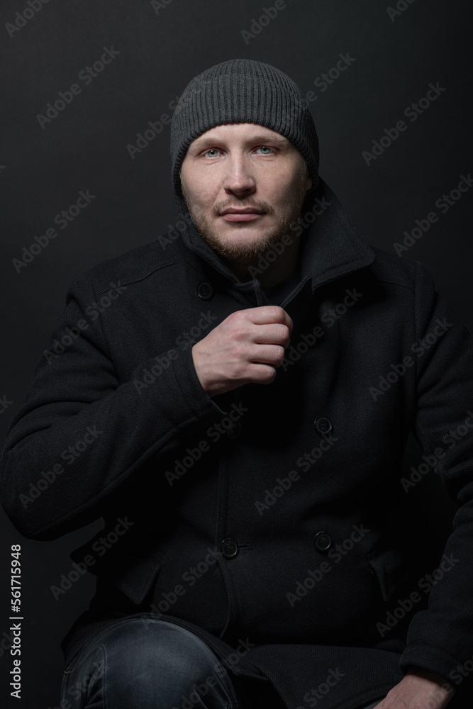 Lifestyle, fashion, occupation concept. Man with winter hat and black coat studio portrait. Model with beard looking at camera with serious look. Dark studio background Lifestyle, fashion, occupation concept. Man with winter hat and black coat studio portrait. Model with beard looking at camera with serious look. Dark studio background