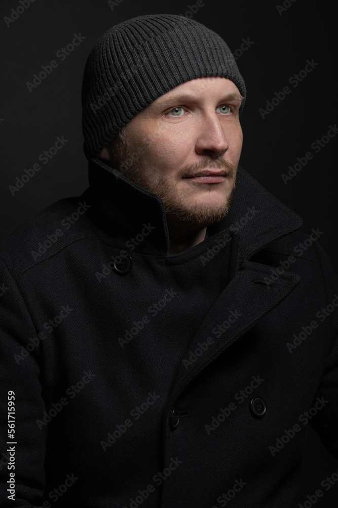 Lifestyle, fashion, occupation concept. Man with winter hat and black coat studio portrait. Model with beard looking aside the camera with serious look. Dark studio background Lifestyle, fashion, occupation concept. Man with winter hat and black coat studio portrait. Model with beard looking aside the camera with serious look. Dark studio background