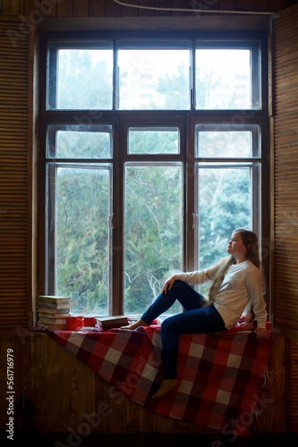 Charming young dreamy girl sits on a window with a book and cup and thinks about something. Concept of coziness and autumn © Fotoproff