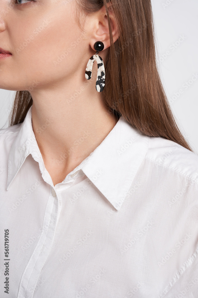 Cropped portrait of a young brown-haired lady in white clothes. The pretty girl is wearing black, white and silver stud earrings with pendants made as a horseshoe and posing on the light background.