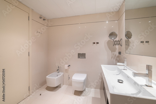 Large bathroom decorated with beige tiles. Square full length mirror and rectangular sink with two faucets  toilet and bidet.