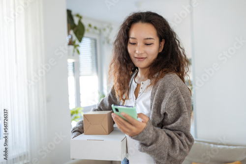 Fotótapéta Smiling young Asian woman shopping online with smartphone on hand, receiving a delivered parcel by home delivery service