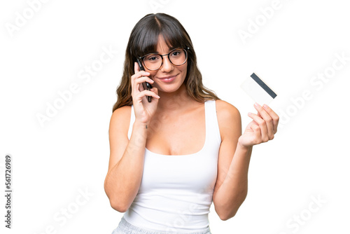 Young caucasian woman over isolated background keeping a conversation with the mobile phone and holding a credit card