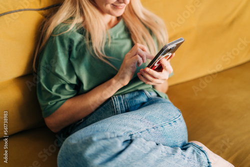 Slika na platnu Unrecognizable woman browsing media on mobile phone while sitting on yellow sofa at home