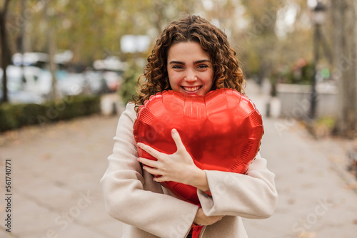 Happy young woman hugging red heart shaped balloon at footpath