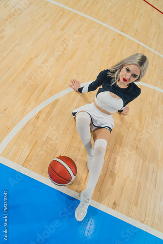 High angle shot of a blond cheerleader sitting on the floor with her hands put also on the floor and a basketball in front of her. Sport concept. Vertical photo. High quality photo