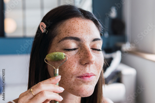 Young woman with freckle face massaging using jade stone roller at home photo