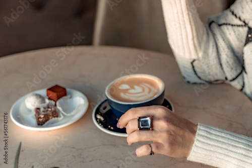 A cup of fresh cappuccino coffee in the hands of a woman on a fashionable background of a white marble table, next to a plate with sweets. Coffee addiction