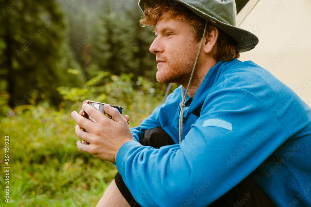 Young white tourist man drinking tea while sitting at tent in forest Young white tourist man drinking tea while sitting at tent in forest