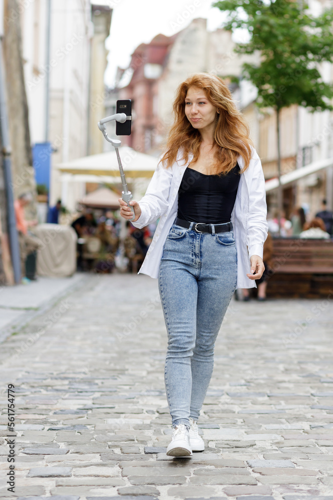Young redheaded beautiful woman walking through the old city and using her mobile phone for video blogging. Pretty smiling girl chatting online