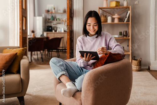 Young smiling asian woman drawing on digital tablet with stylus while sitting in living room