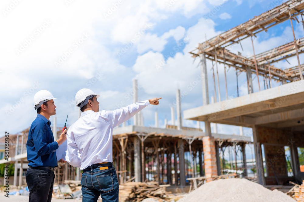 Two engineers are working together and planning in a construction site environment. for architectural business and pointing to the steel structure wearing a helmet and in his hand a radio.