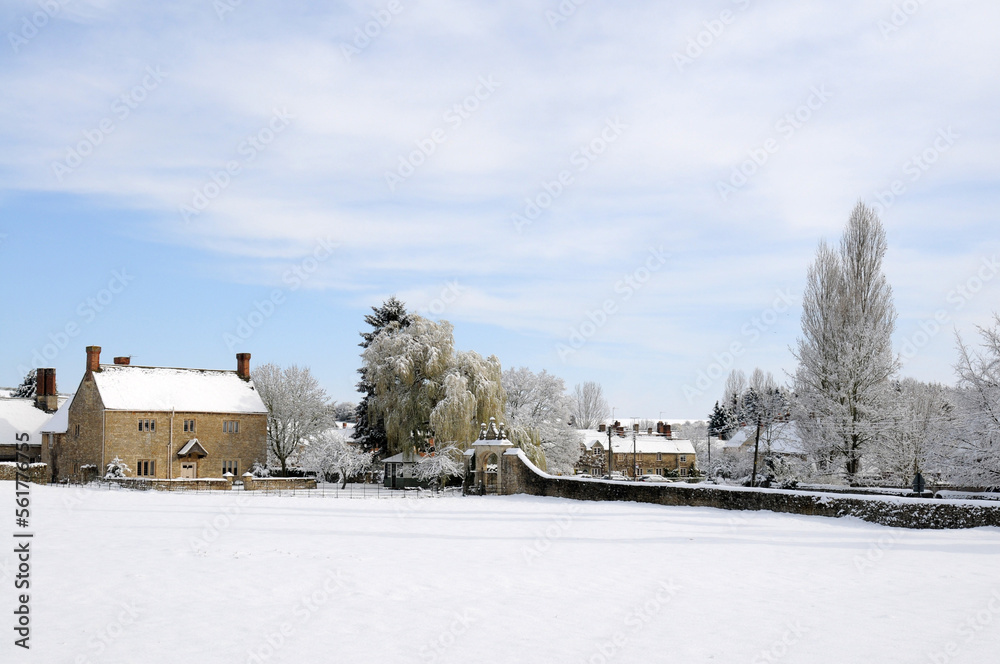 Manor house in the Cotswolds covered in snow with a village behind,  Blue sky and clouds.  poplar trees and a willow tree.