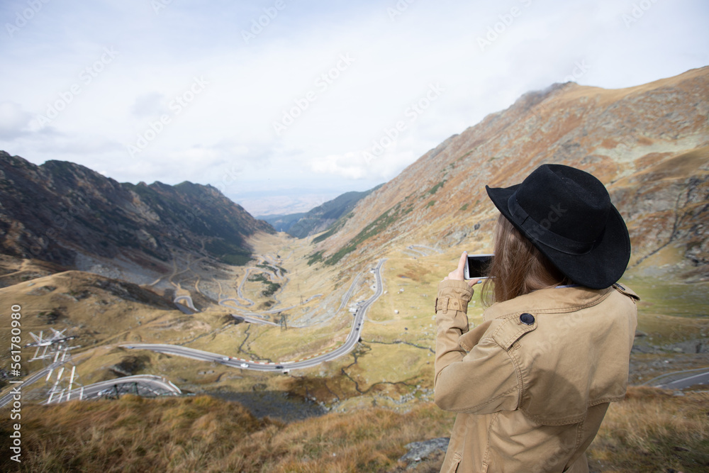 a girl in a raincoat and a black hat takes a photo of a panorama in the mountains on her phone. the girl turns her back to the camera and films the serpentine road on her phone. a girl in a raincoat and a black hat takes a photo of a panorama in the mountains on her phone. the girl turns her back to the camera and films the serpentine road on her phone.