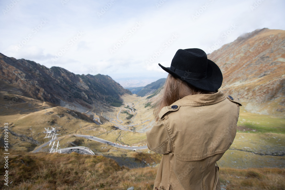 a girl in a black hat stands and looks at the mountains and the serpentine road. Transfagaras Mountains, Romania