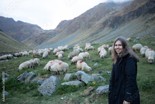 a girl with a big dog sit on the grass in the mountains and watch the sheep. a woman tourist with a shepherd dog herds sheep. evening