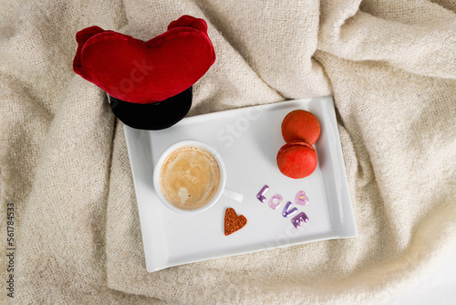 Tray with cup of coffee in bed and macaroni cookies with blanket, love lettering and red heart, breakfast for valentine's day seen from above