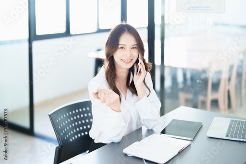 Portrait of a young Asian woman showing a smiling face as she uses his phone, computer and financial documents on her desk in the early morning hours © Jirapong