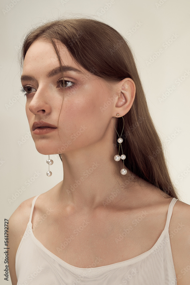 Cropped close-up shot of a young woman with gold stud earrings decorated with pearl beads. A girl with pearl beaded earrings is on a pastel background. Front view.