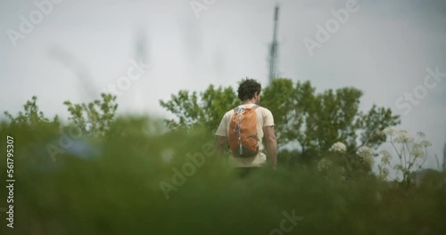 Hiker with an orange backpack walkig toweards the radio tower on mountain Slavnik, camera behind greenery on low perspective. Radio tower visible behind the trees. photo