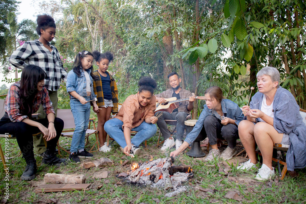 Happy friends group doing recreation with man playing guitar while friends and children grill marshmallow at camping together in holiday, tourist and picnic, party bonfire in vacation. Happy friends group doing recreation with man playing guitar while friends and children grill marshmallow at camping together in holiday, tourist and picnic, party bonfire in vacation.