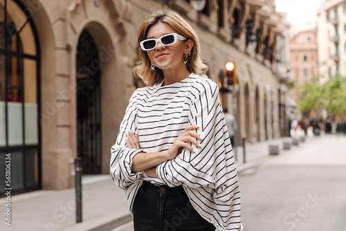 Beautiful caucasian woman look at camera in sunglasses. Young blonde keep crossed hands on the street wearing striped shirt. Streetstyle concept