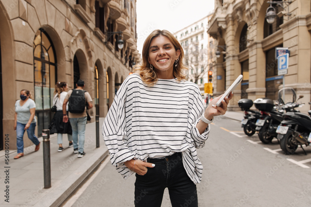 Image of stylish young woman looking with smile at camera staying on the street. Caucasian blonde wearing striped shirt in hands phone. Street style concept