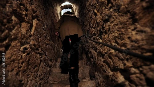 a girl in a raincoat climbs the stairs along a narrow corridor in the fortress. steps of a girl on the catacomb stairs. tunnel in the castle. general plan