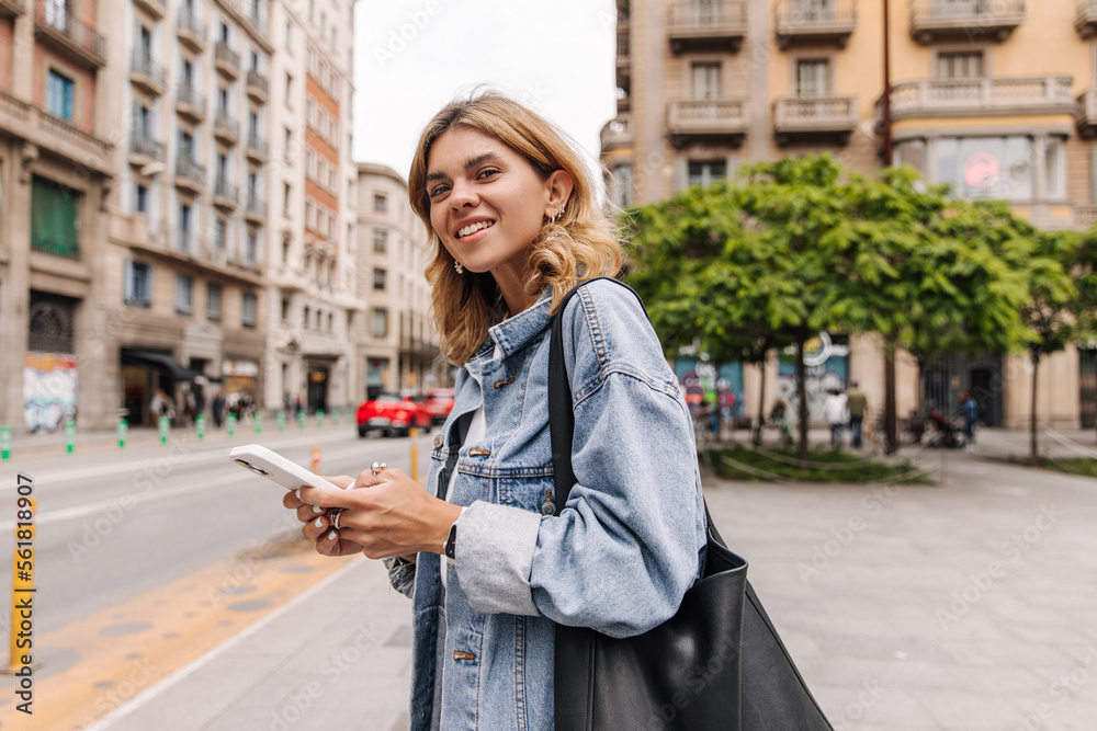 Fototapeta premium The side view of beautiful european woman looking at camera with smile on the street. Fair-skinned blonde wearing jacket holding phone. Street style concept