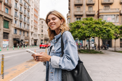 The side view of beautiful european woman looking at camera with smile on the street. Fair-skinned blonde wearing jacket holding phone. Street style concept