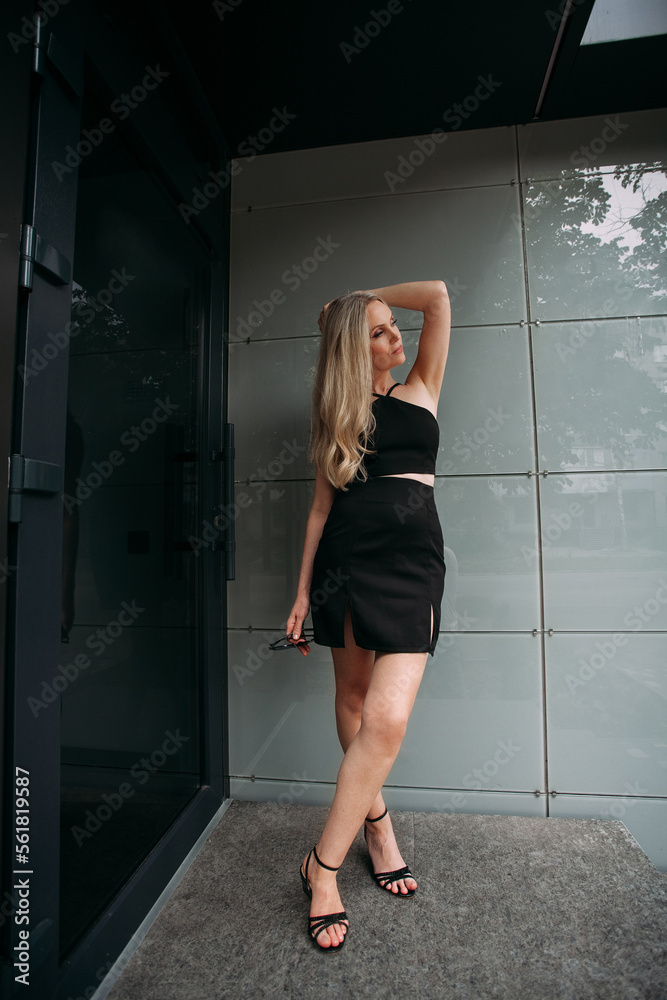 A stylish slender girl with blond long hair and long legs stands against the backdrop of a modern building. Dressed in a short black sexy skirt and top. A stylish slender girl with blond long hair and long legs stands against the backdrop of a modern building. Dressed in a short black sexy skirt and top.