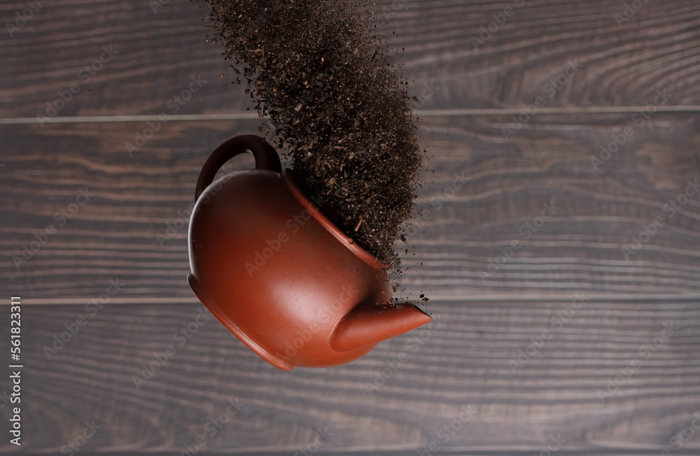 Fototapeta premium Black dried leave Tea fly in Pot. Small Fine size tea leaf flying explosion, Abstract cloud fly. Brown colored Teas pot splash throwing in Air. Wooden background Isolated high speed freeze