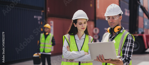 Cheerful Caucasian engineer wearing safety vest standing by shipping container terminal talking discussing information on computer to collaborator. Transportation business. Logistic industrial concept
