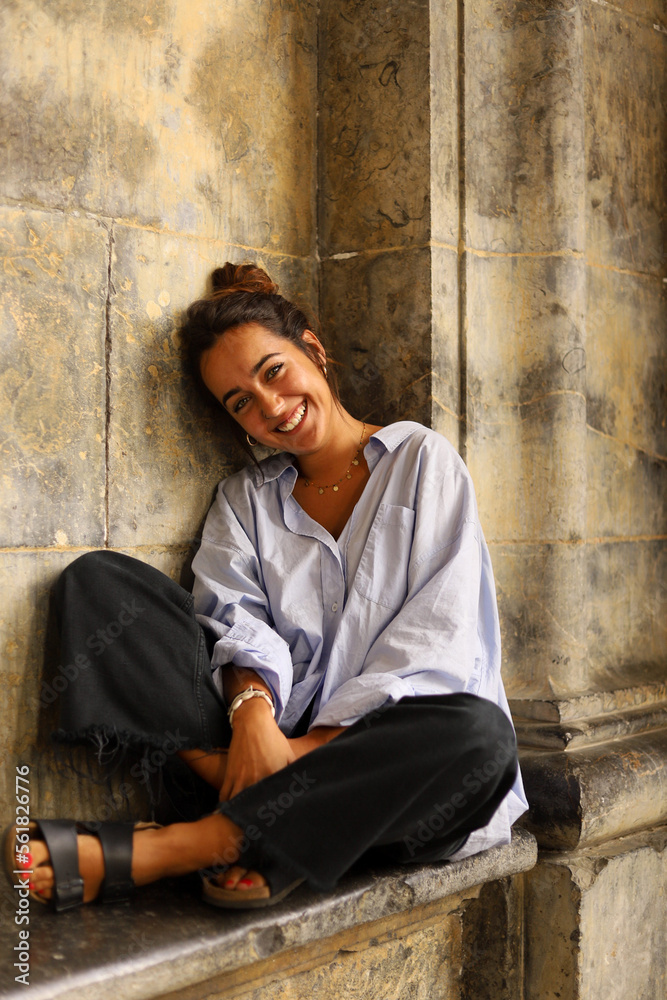 Smiling young lady sitting on stone bench near aged building and looking at camera Smiling young lady sitting on stone bench near aged building and looking at camera