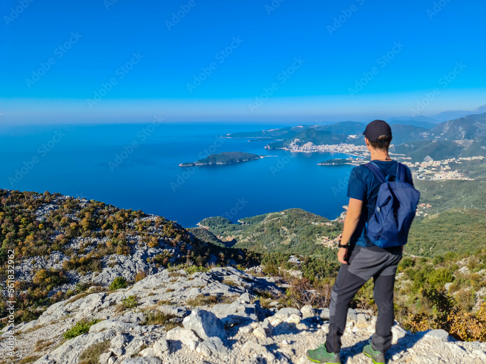 Hiking man with backpack enjoying panoramic aerial view on coastline of Budva and Sveti Nikola Island seen from Goli Vrh, Adriatic Mediterranean Sea, Montenegro, Balkan, Europe. Budvanian Riviera